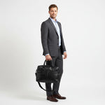 Chris Louis black leather briefcase with detachable shoulder strap, held by a woman in a white shirt and grey trousers.