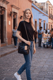 A woman wearing a Chris Louis black leather satchel bag with a drawstring closure while walking on a cobblestone street.