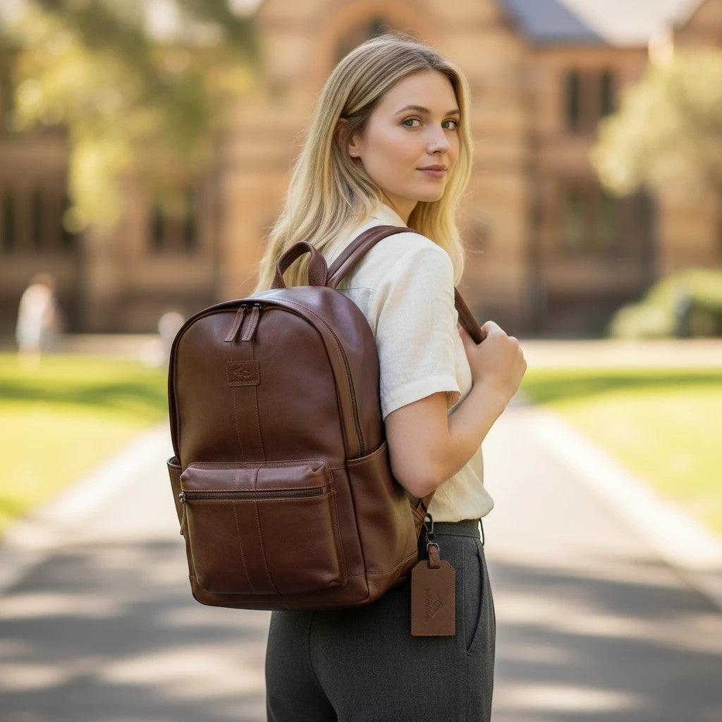 Chris Louis warm brown genuine leather backpack with front zip pocket, side pockets, and detachable luggage tag. Model wears it with grey trousers.