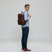Chris Louis deep brown genuine leather backpack with multiple pockets and padded straps, worn by a woman on a cobblestone street.