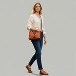 Chris Louis Santorini tan leather crossbody bag with front flap and metal hardware, worn by a woman walking on a cobblestone street.