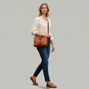 Chris Louis Santorini tan leather crossbody bag with front flap and metal hardware, worn by a woman walking on a cobblestone street.