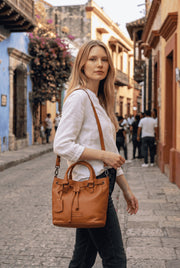 A woman wearing a Chris Louis tan full grain leather satchel bag with drawstring and crossbody strap on a city street.